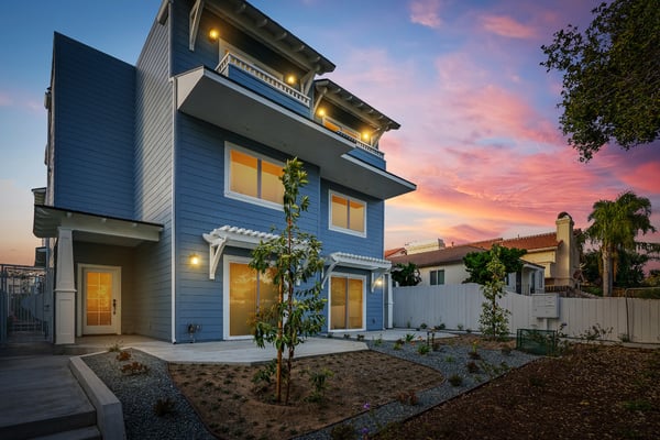 Modern blue two-story house with lit balconies, concrete patio, young landscaping, and sunset sky at dusk