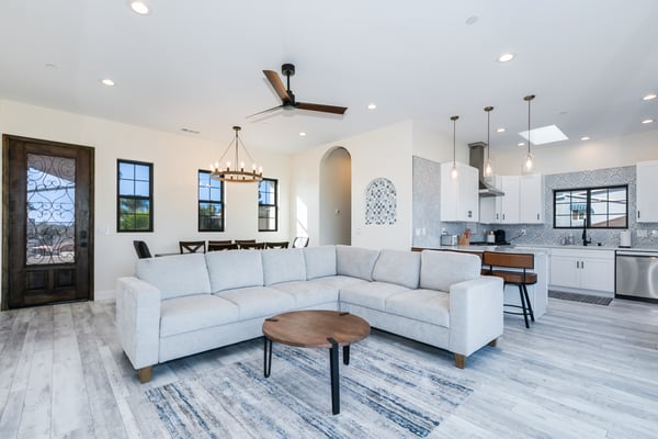 Open-plan living room with light blue sectional sofa, wood coffee table, and kitchen with white cabinetry and mosaic tile backsplash