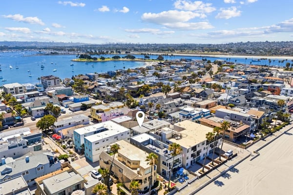 Aerial view of coastal neighborhood with residential homes, palm trees, sandy beach, and blue bay with sailboats