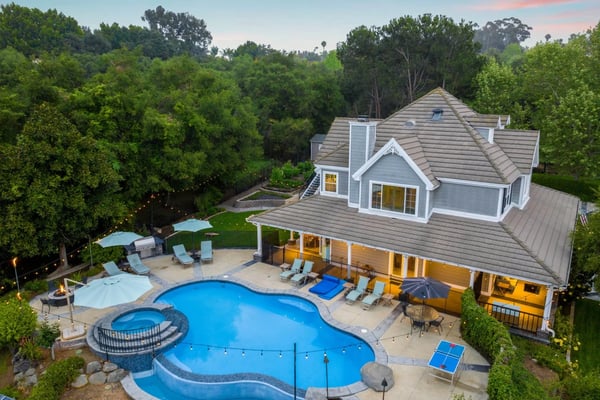 Modern home with curved swimming pool, spa, lounge chairs, and covered patio surrounded by mature trees at sunset.