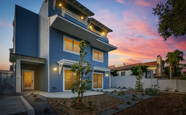 Modern blue two-story house with lit balconies, concrete patio, young landscaping, and sunset sky at dusk